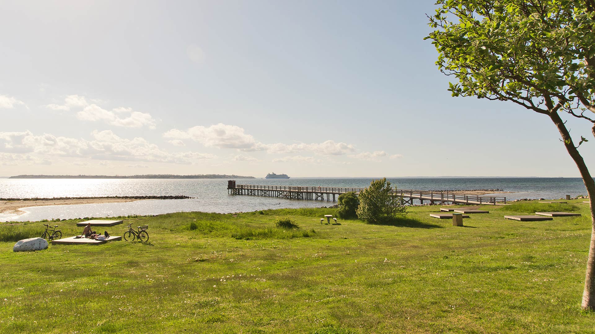 Badplats med brygga vid Öresund med utsikt mot ön Ven och passerande kryssningsfartyg i bakgrunden.